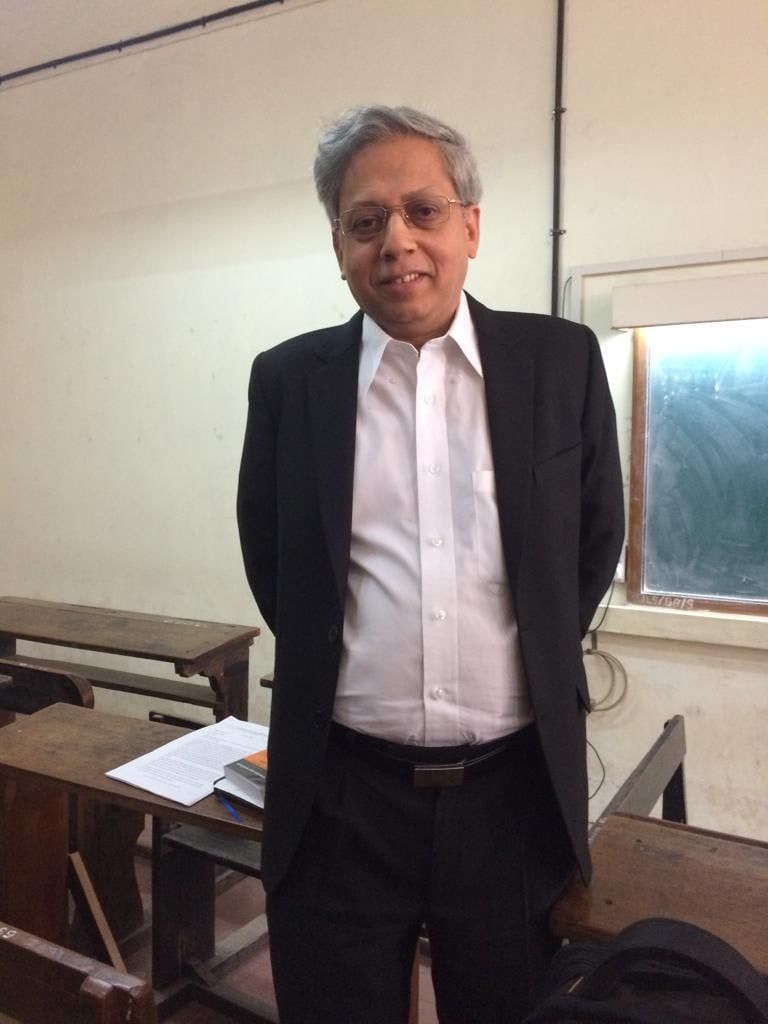 Neelakanth Phadke standing in a classroom of wooden desks, beside an open book and lecture notes.